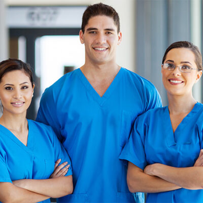 Three healthcare professionals wearing blue scrubs stand together indoors, smiling confidently at the camera. Two women and one man, one of the women wears glasses, and all appear friendly and approachable.