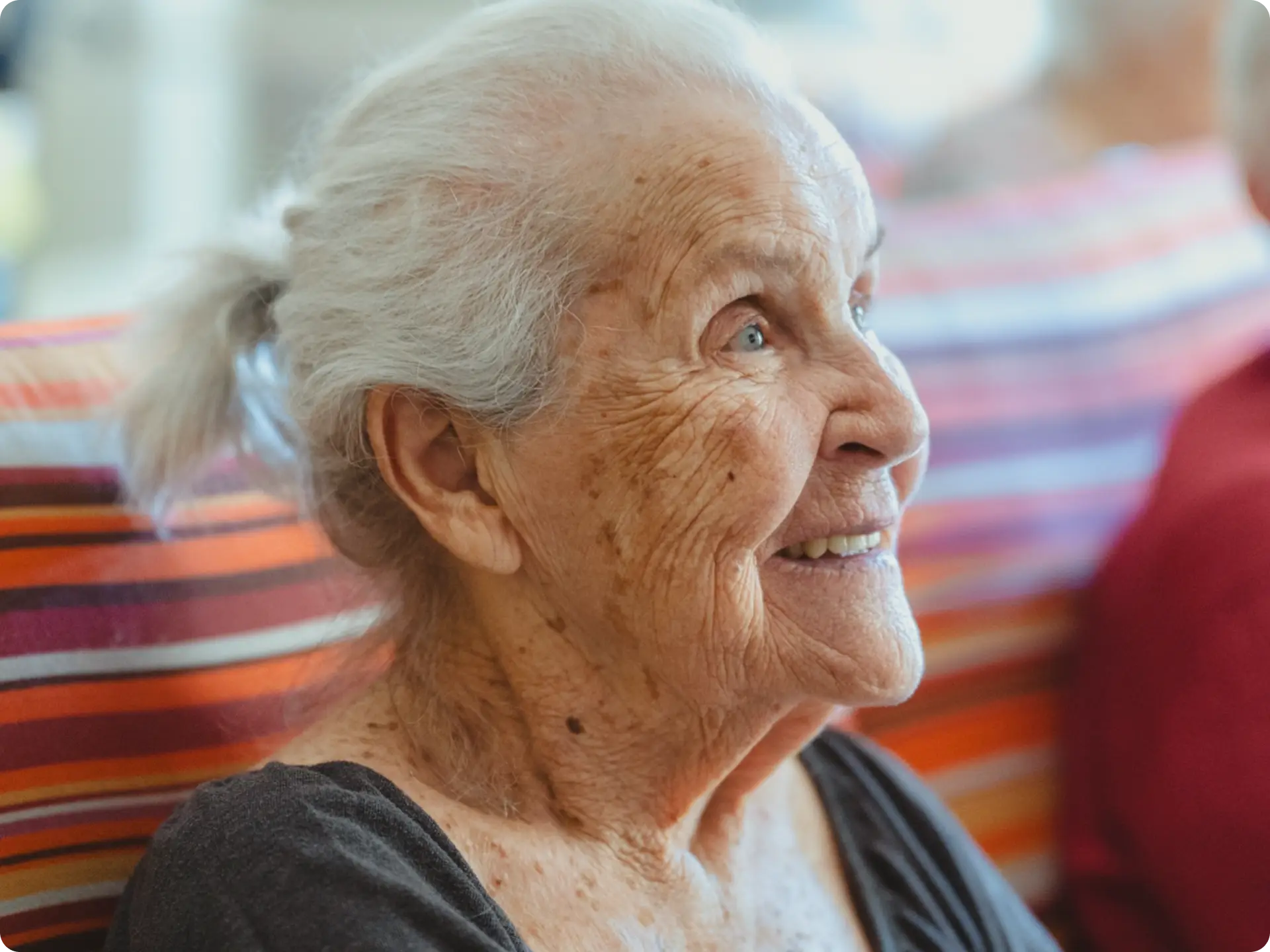 An elderly woman with white hair in a ponytail smiles warmly while sitting on a colorful striped cushion, looking slightly to the right. She wears a dark top and appears content and relaxed.