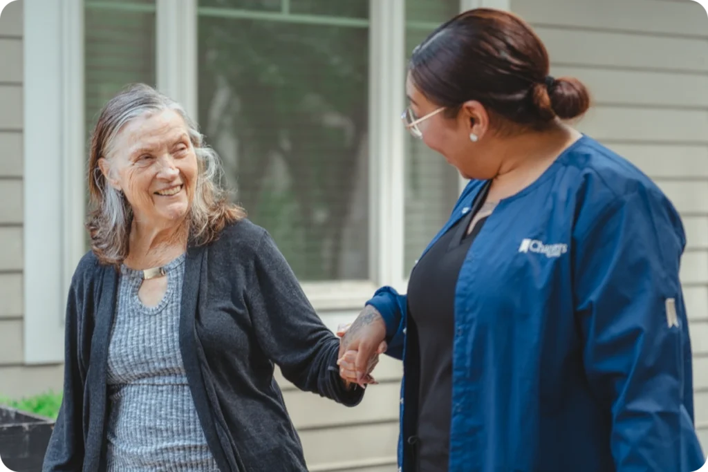An elderly woman smiles while holding hands with a caregiver in a blue uniform as they walk outside near a house. The caregiver looks at her kindly, and they both appear happy and engaged.