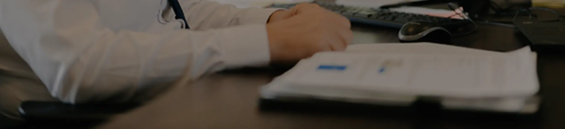 A person in a white shirt sits at a desk with folded hands near a computer keyboard, mouse, and an open notebook or document.